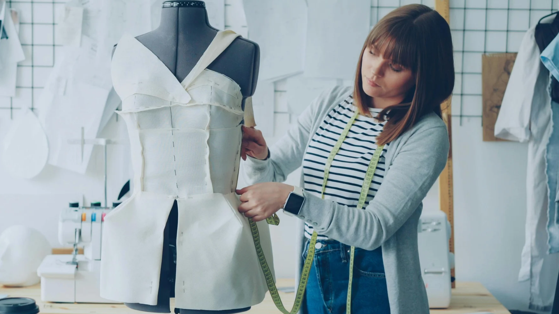 Fashion designer adjusts a pinned cream fabric garment on a dress form, with a measuring tape around their neck.
